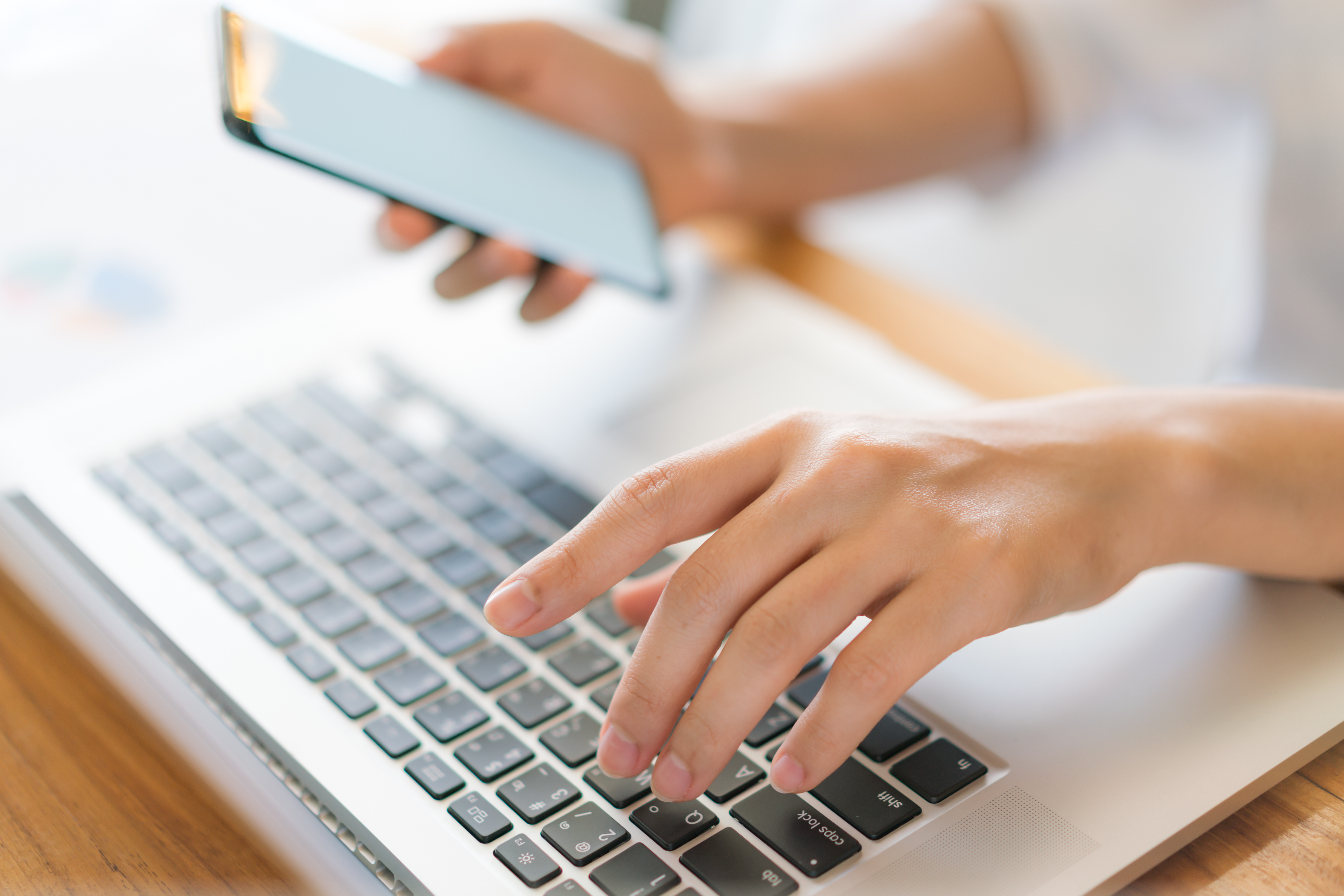 Osoba siedząca przy laptopie oraz&nbsp;trzymająca w&nbsp;dłoni telefon kom&oacute;rkowy