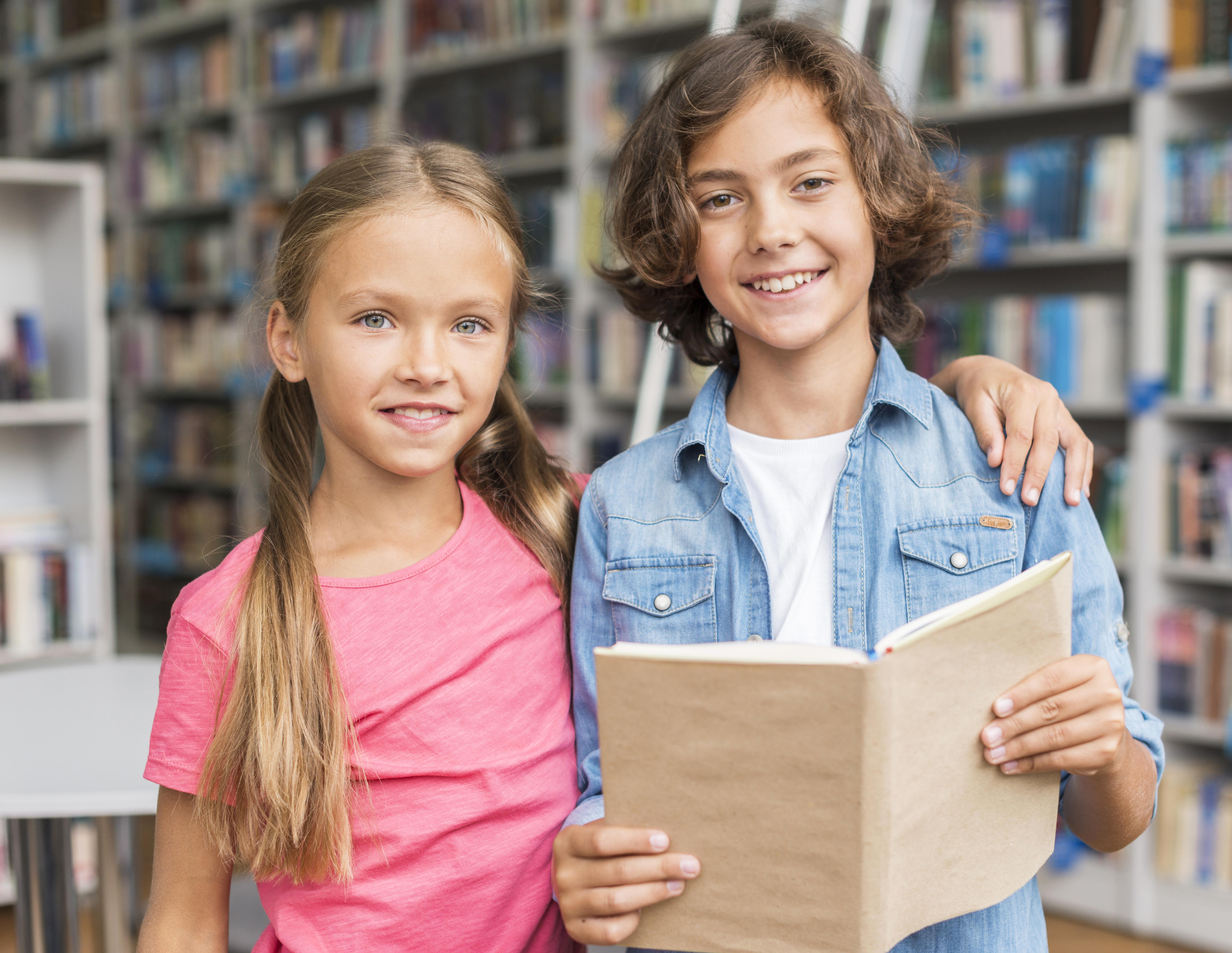 Dziewczyna i&nbsp;chłopiec. Chłopiec trzyma książkę. Za nimi regały z&nbsp;książkami. Prawdopodobnie są w&nbsp;Bibliotece